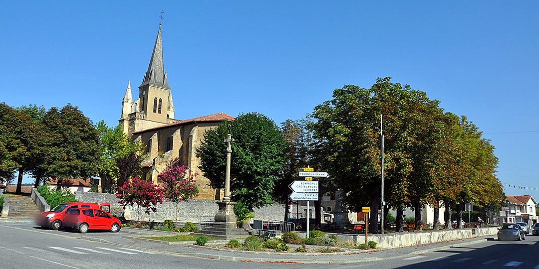 L'église de Galan et son clocher en pierre sous un ciel bleu, avec une croix de pierre et une allée d'arbres.